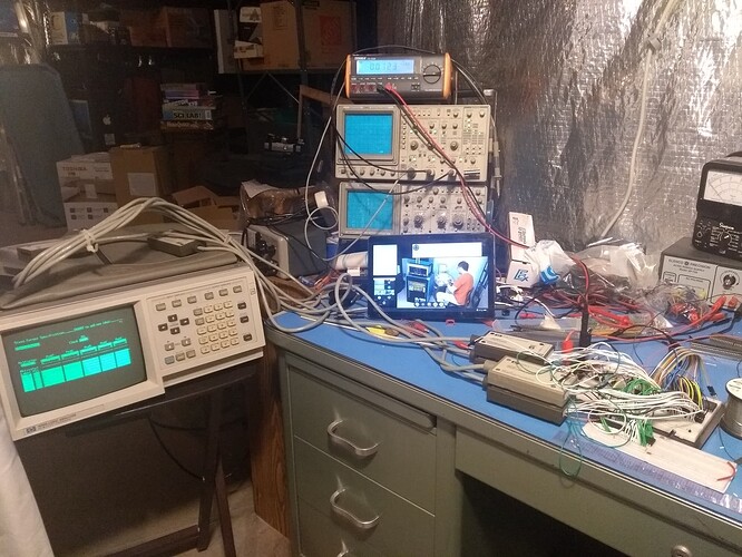 A very messy green tanker desk with the homebrew in progress on its surface on the right, and a cluttered unfinished basement in the background.  A logic analyzer is visible on a small table to the left, and a stack of two oscilloscopes and a DMM sits above a tablet playing a video of someone operating a PDP-8 on the desk  The desk has a variety of other equipment on it, such as rolls of solder and a Simpson VOM.  The wall behind the desk is covered with foil-backed insulation.