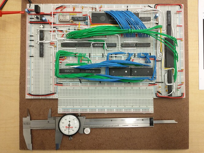 A much neater set of solderless breadboards than the previous pictures is squared nicely in the frame affixed to a brown hardboard backer with a Starrett dial caliper below.  There are four breadboards in horizontal orientation down the center, with a vertical breadboard on either side.  Neat wiring criscrosses the boards in square-cornered traces and smooth bundles forming arches.  The bundles of wires are mostly mono-chromatic in either blue, green, or white.  Chips are laid densely on four of the boards, loosely on the left vertical board, and the lowest horizontal breadboard is empty.
