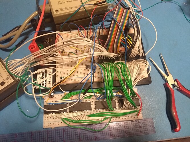 A double solderless breadboard on a backing plate sits on a blue ESD mat with a mess of wires connecting it to a third solderless breadboard below.There is only a single IC on the center breadboard, which appears to be used as mostly for signal distribution.  A variety of wires and chips populate the boards, with wiring in various colors that looks intentional in some places.Logic analyzer pods are connected to the center breadboard with flying leads.  A craft ruler and a pair of needlenose pliers sit on the desk beside the breadboards.