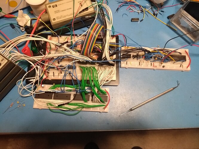 A double solderless breadboard wired loosely to a second solderless breadboard below and a third solderless breadboard to the right sit on a blue ESD mat.  There are five logic analyzer pods scattered near it with flying leads probing a number of different points.  A few DIP ICs, some resistors and capacitors, a dental pick, and assorted detritus surround the breadboards.  A variety of wires and chips are installed on the boards.