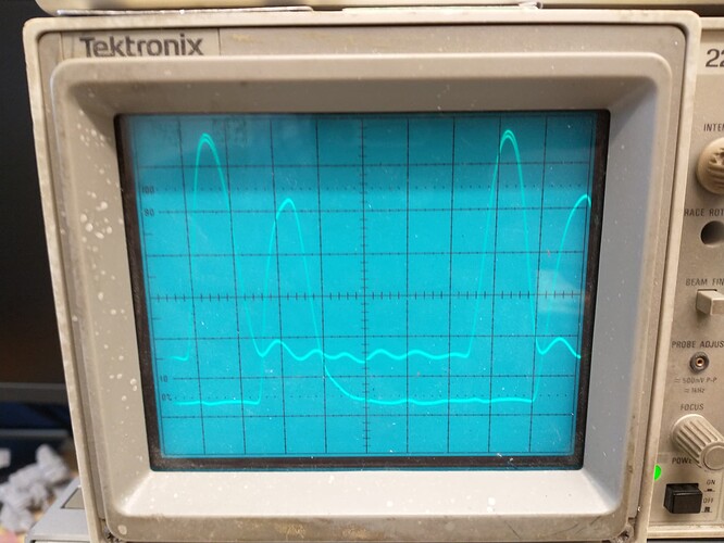 A grungy Tektronix oscilloscope CRT screen showing two traces.  The upper trace shows two periods of about 1/4 duty cycle of either a very rounded square wave or the positive going portion of a sloppy sine wave, while the lower trace shows two periods of similar shape approximately 1/4 cycle later.