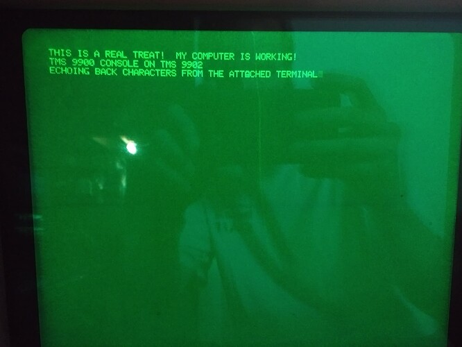 A glowing green CRT with the text "This is a real treat!  My computer is working!TMS 9900 console on TMS 9902Echoing back characters from the attached terminal" in a bitmapped font in all capital letters.  The second A in attached is visibly overstruck with some other character.  There is a reflection in the glass of someone taking a photo with a phone.